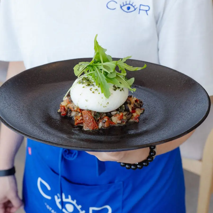 A COR server presenting a burrata dish on a black plate
